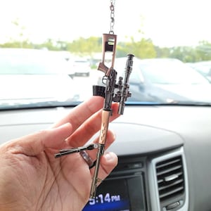 May include: A black and silver miniature rifle with a scope hanging from a rearview mirror in a car. The rifle has a brown wooden stock and a black metal barrel. The rifle is a popular collectible and is often used as a car charm.