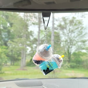May include: Two clear plastic cups with a black and white cat design hanging from a rearview mirror. The cups are filled with a pink and blue liquid and black boba balls. The text on the cups reads "Cat Milk Cap Drinks".