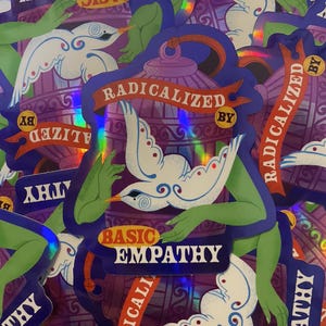 Peut inclure: Plusieurs autocollants holographiques avec un design violet et bleu. Les autocollants présentent un oiseau aux ailes déployées, des bras verts et le texte "RADICALIZED BY BASIC EMPATHY".