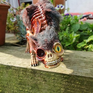 May include: A skeletal cat figurine with a red and brown body, a grey fur coat, and one glowing green eye. The figurine is posed in a walking position with its head tilted down.