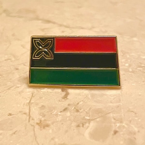 May include: A gold-colored pin with a red, black, and green flag design. The flag features a stylized symbol in the top left corner.