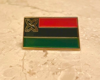 May include: A gold-colored pin with a red, black, and green flag design. The flag features a stylized symbol in the top left corner.