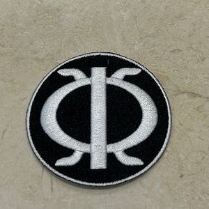 May include: A black and white embroidered patch with a stylized symbol. The symbol is a circle with a vertical line in the center and a curved line on each side of the line.