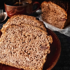 May include: Close-up of a sliced loaf of brown bread on a brown plate. The bread has a crumbly texture and is slightly darker in color on the edges.