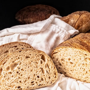 May include: Close-up of three loaves of freshly baked bread, two of which are sliced open, revealing a soft, airy crumb. The bread is golden brown and has a rustic appearance.