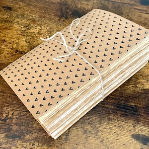 May include: A stack of rectangular paper sheets wrapped in a tan paper cover featuring a repeating pattern of small black hearts. The stack is secured with a thin, light-colored twine tied in a bow across the top of the paper cover.