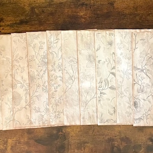 May include: A collection of rectangular placemats with a floral pattern in shades of cream and gray. The placemats are arranged in a row on a dark wooden surface, showcasing their delicate design.