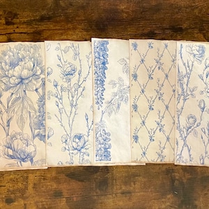 May include: A collection of folded paper sheets with blue floral patterns on a cream background. The designs include peonies, roses, and other botanical elements. The papers are arranged on a dark wooden surface.