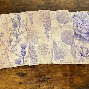 May include: A set of six off-white fabric napkins with blue floral designs. Each napkin features a different botanical illustration, including peonies, thistles, and wisteria. The napkins are arranged on a wooden surface.