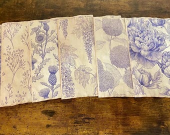 Puede incluir: Un juego de seis servilletas de tela blanquecina con diseños florales azules. Cada servilleta presenta una ilustración botánica diferente, incluyendo peonías, cardos y glicinias. Las servilletas están dispuestas sobre una superficie de madera.