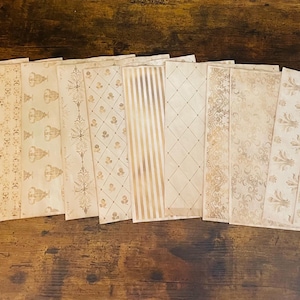 May include: A collection of decorative paper sheets with various vintage-style patterns. The sheets feature designs such as floral, geometric, and striped motifs in shades of beige and brown. The papers are arranged on a dark wooden surface.