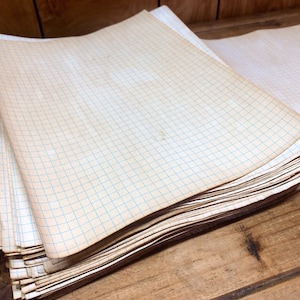 May include: A stack of blank, white graph paper with blue grid lines. The paper is slightly yellowed with age and has a worn, vintage look.