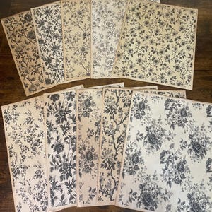 May include: A collection of vintage-style paper sheets with a floral pattern. The design features black and gray flowers and foliage on an off-white background. The sheets are arranged on a dark wooden surface.