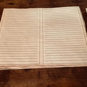May include: A stack of lined paper sheets with a black border and lines. The paper has a vintage, aged appearance, with a slightly yellowed hue. The sheets are stacked on a dark brown wooden surface.