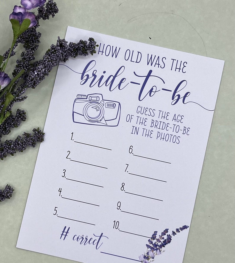 May include: A white card with purple text and a purple camera illustration. The text reads 'How old was the bride-to-be? Guess the age of the bride-to-be in the photos.' There are ten lines with numbers 1 through 10 and blank spaces for answers. The bottom of the card reads '# correct' with a blank space for the answer. There are purple flowers in the bottom right corner of the card.