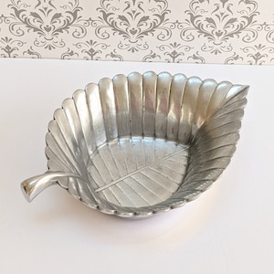 Puede incluir: Un plato de servir plateado en forma de hoja con una superficie texturizada. El plato tiene un asa en el lateral.