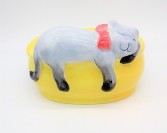 May include: A ceramic figurine of a gray cat with a pink bow, sleeping on a yellow oval-shaped bed.