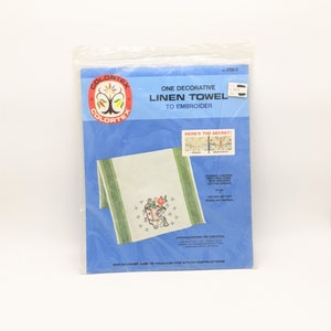 May include: A package containing a white linen towel with a green border and a colorful embroidered design of a birdhouse with flowers. The package includes instructions for embroidery. The text on the package reads "One Decorative Linen Towel to Embroider".