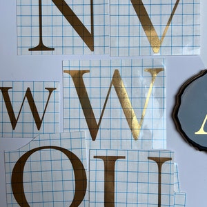 May include: Gold letters on a white gridded background, spelling out the word "NOW" with the letters "U" and "W" repeated.