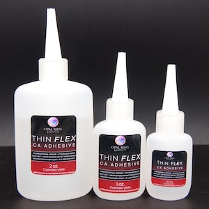 May include: Three white plastic bottles of Thin Flex CA Adhesive, with white spouts and black and red labels. The bottles are labeled with sizes: 2 oz, 1 oz, and 0.5 oz. The labels include the text "Cyanoacrylate".
