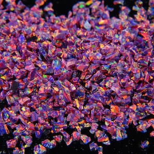 May include: A close-up view of a pile of crushed opal gemstones. The opals are a mix of purple, red, and blue hues, with iridescent flashes of color. The gemstones are irregularly shaped and appear to be rough-cut.