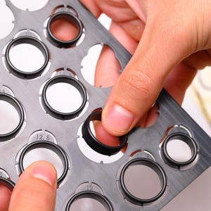 May include: A close-up of a black ring being placed into a gray ring sizer. The sizer has multiple circular openings with size markings. The ring is made of a dark material and has a textured surface.