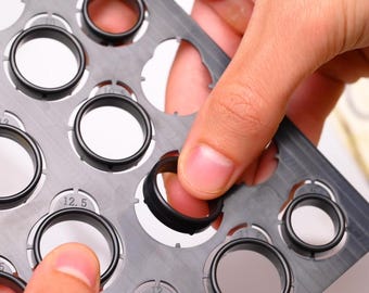 Può includere: Primo piano di un anello nero che viene inserito in un misuratore di anelli grigio. Il misuratore ha più aperture circolari con indicazioni di taglia. L'anello è realizzato in materiale scuro e ha una superficie strutturata.