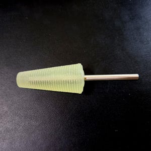 Puede incluir: Una fresa de uñas cónica verde claro con una superficie texturizada y un eje de metal plateado. La broca está diseñada para el cuidado de las uñas y se coloca sobre un fondo oscuro.