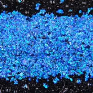 Pode incluir: Uma imagem em close-up de uma pilha de opala azul esmagada e iridescente. As peças de opala são pequenas e de forma irregular, com uma qualidade brilhante e reflexiva. O fundo é uma superfície escura e molhada.