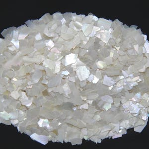 Könnte beinhalten: Ein Haufen schillernder, weißer Muschelsplitter. Die Flocken sind unregelmäßig geformt und haben einen perlmuttartigen Glanz, der das Licht reflektiert. Das Material ist wahrscheinlich für dekorative Zwecke gedacht.