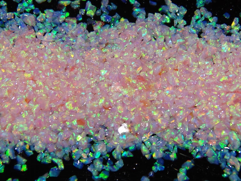 May include: A close-up of a pile of iridescent pink and green opal chips. The chips are small and irregular in shape, and they shimmer and sparkle in the light.