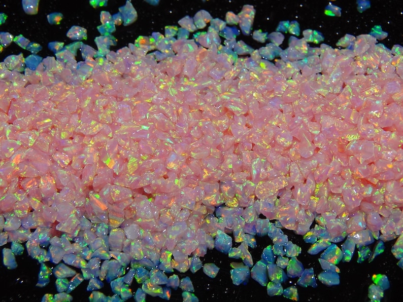 May include: A close-up of pink and iridescent opal chips. The chips are scattered on a black surface.
