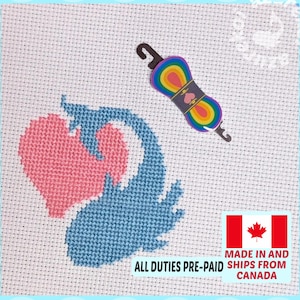 May include: A cross-stitch pattern on white Aida fabric featuring a pink heart and a blue whale, accompanied by a rainbow-colored embroidery floss skein on a black plastic bobbin. The design displays a whale shape composed of a pink heart and blue body.