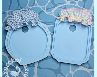 May include: Two light blue silicone molds, one circular and one rectangular, each featuring a central hole and a fabric cover. The circular mold has a blue and white gingham cover, while the rectangular mold has a pastel plaid cover.
