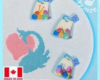 May include: Three tea bag-shaped needle minders featuring colorful yarn balls and crafting tools, placed on white Aida fabric with a cross-stitched pink heart and blue whale design. Each tag is labeled Craft Tea with a black string and metal hook.
