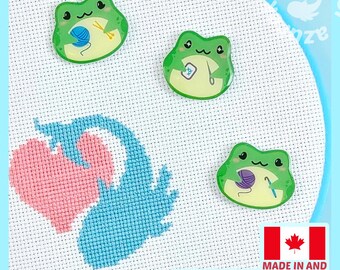 May include: Three green frog-shaped needle minders with glossy finishes, each depicting a frog engaged in a different craft activity, placed on a white Aida fabric square featuring a cross-stitched design of a pink heart and a blue axolotl.