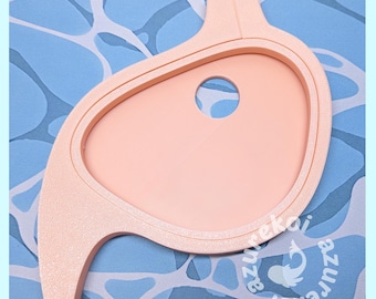 May include: A peach-colored, asymmetrical, organic-shaped plastic cookie cutter with a single circular cutout in the center. The surface has a textured finish, and the design features a curved, tapered tail-like extension on one side.
