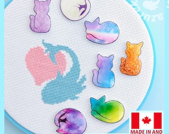 May include: A circular embroidery hoop containing white Aida fabric with a cross-stitched pink heart and blue whale design. Seven cat-shaped needle minders with various colorful patterns, including galaxy, rainbow, and solid hues, are attached.