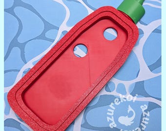 May include: A red, rectangular, 3D-printed frame shaped like a hot sauce bottle with a green cap at the top. The frame features two circular cutouts in the center and a textured surface. The text "azurekoi" is visible in the lower right corner.