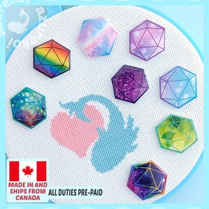May include: Seven multi-coloured polyhedral dice-shaped pins arranged on a white cross-stitch fabric background. The pins feature various designs including rainbow, galaxy, floral, and glitter patterns with black geometric line overlays.