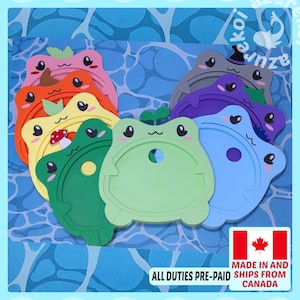 May include: A collection of colorful, flat, frog-shaped plastic frames in shades of pink, orange, yellow, green, blue, purple, and grey. Each frame features a small sprout on top, a central circular cutout, and a smiling face with large eyes.