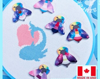 May include: Six bee-shaped needle minders with galaxy-patterned wings and colorful striped bodies arranged on a white Aida fabric cross-stitch project inside a blue embroidery hoop. The bee designs feature various pride flag color combinations.