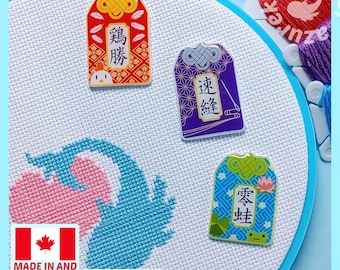 May include: Three rectangular omamori-style charms featuring Japanese characters and illustrations, including a red charm with a chicken, a purple charm with a needle and thread, and a blue charm with a frog, placed on white cross-stitch fabric.