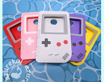May include: A collection of five handheld gaming console cases in red, pink, white, purple, and yellow. Each case features a screen cutout, a directional pad, two circular buttons, and two horizontal start and select buttons on the front.
