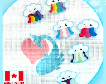May include: Six cloud-shaped enamel pins featuring various rainbow and pride flag color patterns, each with a smiling face. The pins are arranged on a white cross-stitch fabric background that features a stitched pink heart and blue whale design.