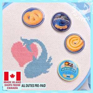 May include: A white cross-stitch fabric featuring a pink heart and blue whale design, accompanied by four circular pins: one with a pretzel, one with a Royal Dansk cookie tin, one with a swirl cookie, and one labelled Sewing Kit with cookie graphics.