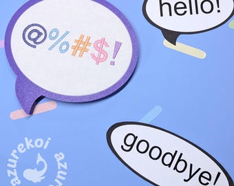 May include: Three speech bubble signs on a blue background. One is a purple-framed embroidery hoop with white fabric featuring the embroidered text @%#$!. Two other white speech bubbles have black outlines and the printed text hello! and goodbye!