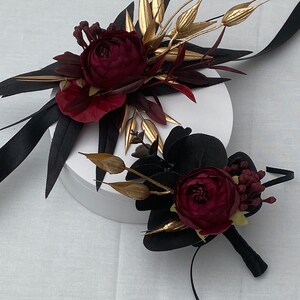 Pu&ograve; includere: Due boutonni&egrave;re floreali di colore bordeaux e oro con accenti di nastro nero. Le boutonni&egrave;re sono su una superficie bianca.