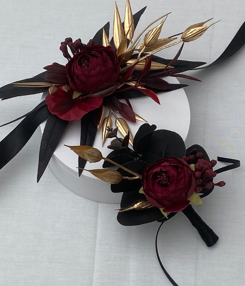 Pu&ograve; includere: Due boutonni&egrave;re floreali di colore bordeaux e oro con accenti di nastro nero. Le boutonni&egrave;re sono su una superficie bianca.
