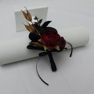 Pu&ograve; includere: Un boutonniere per un matrimonio o un evento speciale con una rosa bordeaux, dettagli dorati e fogliame nero, legato con un nastro nero.